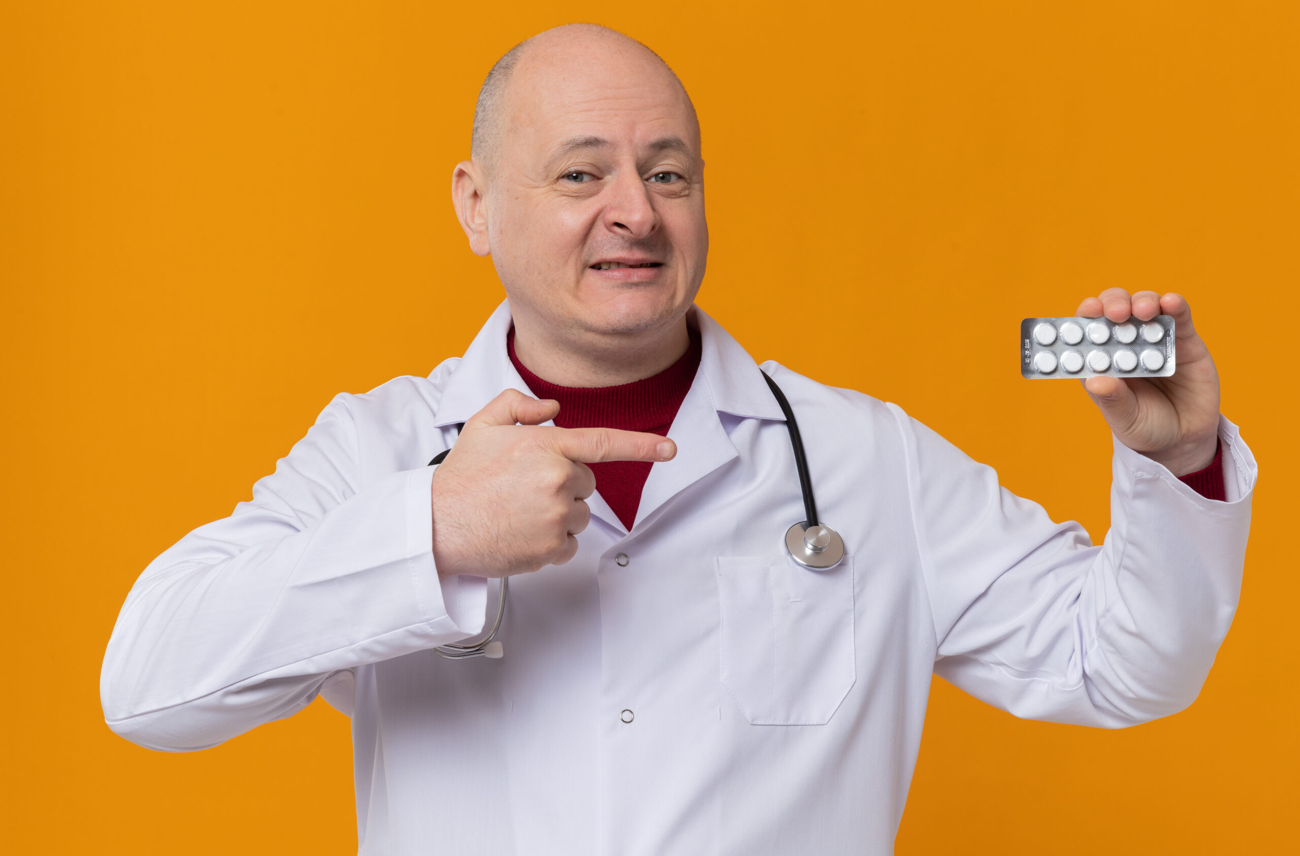 smiling adult slavic man in doctor uniform with stethoscope holds and points at medicine blister pack isolated on orange background with copy space