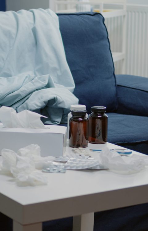 Close up of medicaments and thermometer on coffee table with nobody in room. Empty place with medicine against flu, bottles of pills, drugs, medication, cup of tea and tissues for treatment